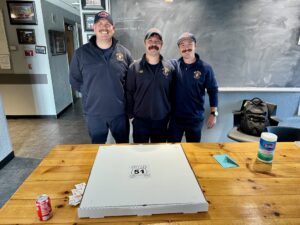three firefighters stand in a row for a picture in front of large pizza - Wallentine Injury Law 2025 Community Involvement Report